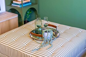 Ein gestreiftes Sofa mit einem Tablett, auf dem ein Glas und eine Flasche stehen, eine kleine Topfpflanze und ein Buch auf einem Regal.