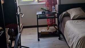 Makuuhuone, jossa on sänky, yöpöytä, pitsiverhoilla varustettu ikkuna, koriste-esineillä täytetty hylly ja puulattia.