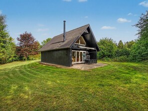 Una casa in legno a un piano con tetto spiovente e un camino, immersa in un prato curato e vegetazione lussureggiante.
