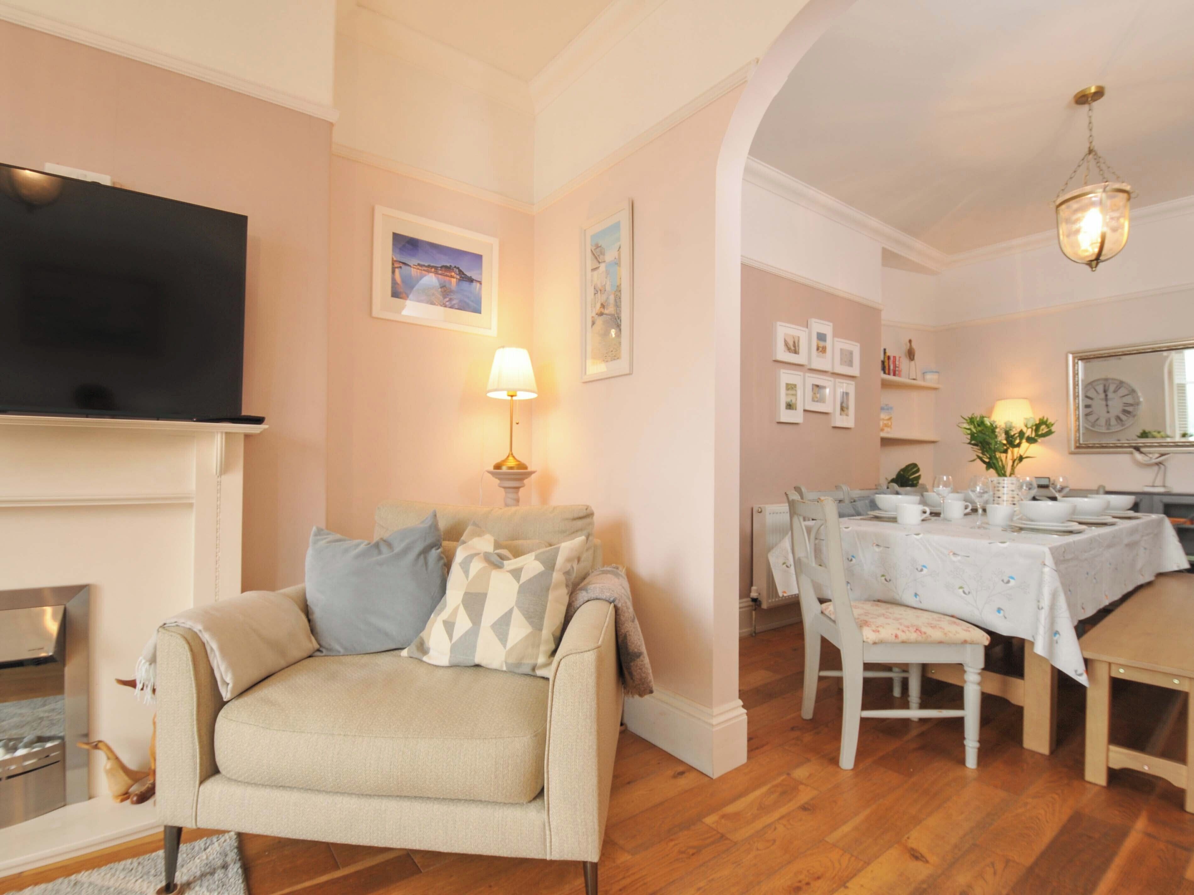 A living room with a sofa, a flat-screen TV, a fireplace, a dining area with a table set for four, and a chandelier.