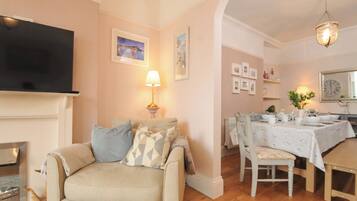 A living room with a sofa, a flat-screen TV, a fireplace, a dining area with a table set for four, and a chandelier.