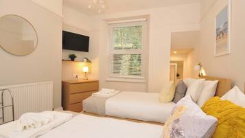 A hotel room with a bed, a bedside table, a mirror, a TV, a window with blinds, and a chandelier.