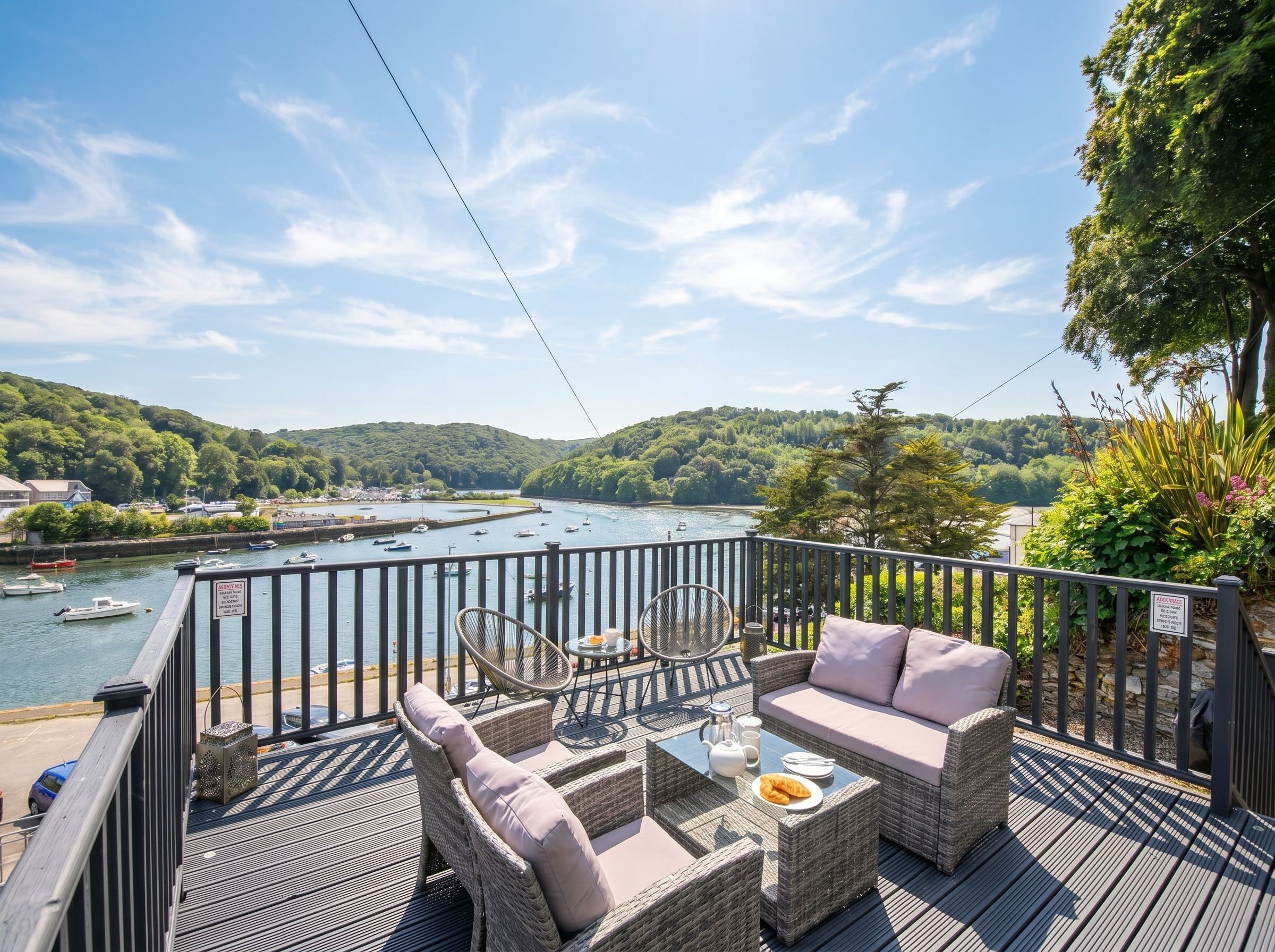 A deck with wicker furniture, a table with a teapot and plate, and a view of a river with boats.