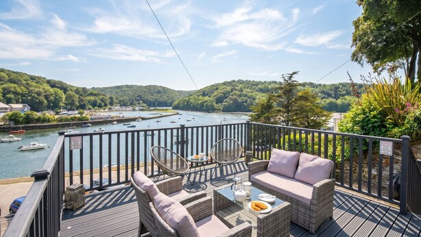 Eine Terrasse mit Korbmöbeln, einem Tisch mit Teekanne und Teller, und Blick auf einen Fluss mit Booten.