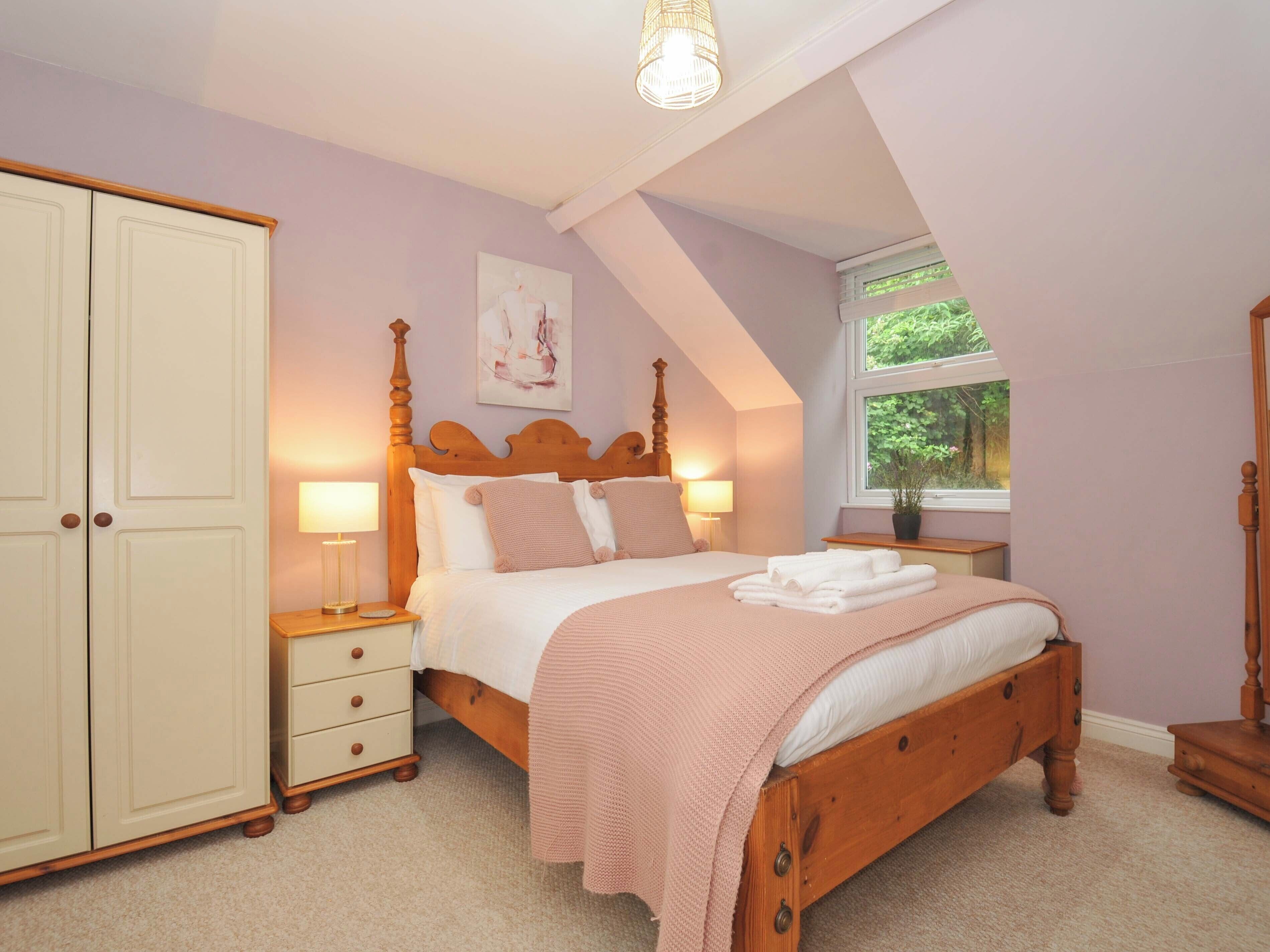 A bedroom with a wooden bed, a nightstand, a dresser, a window with a view of greenery, and a ceiling light.