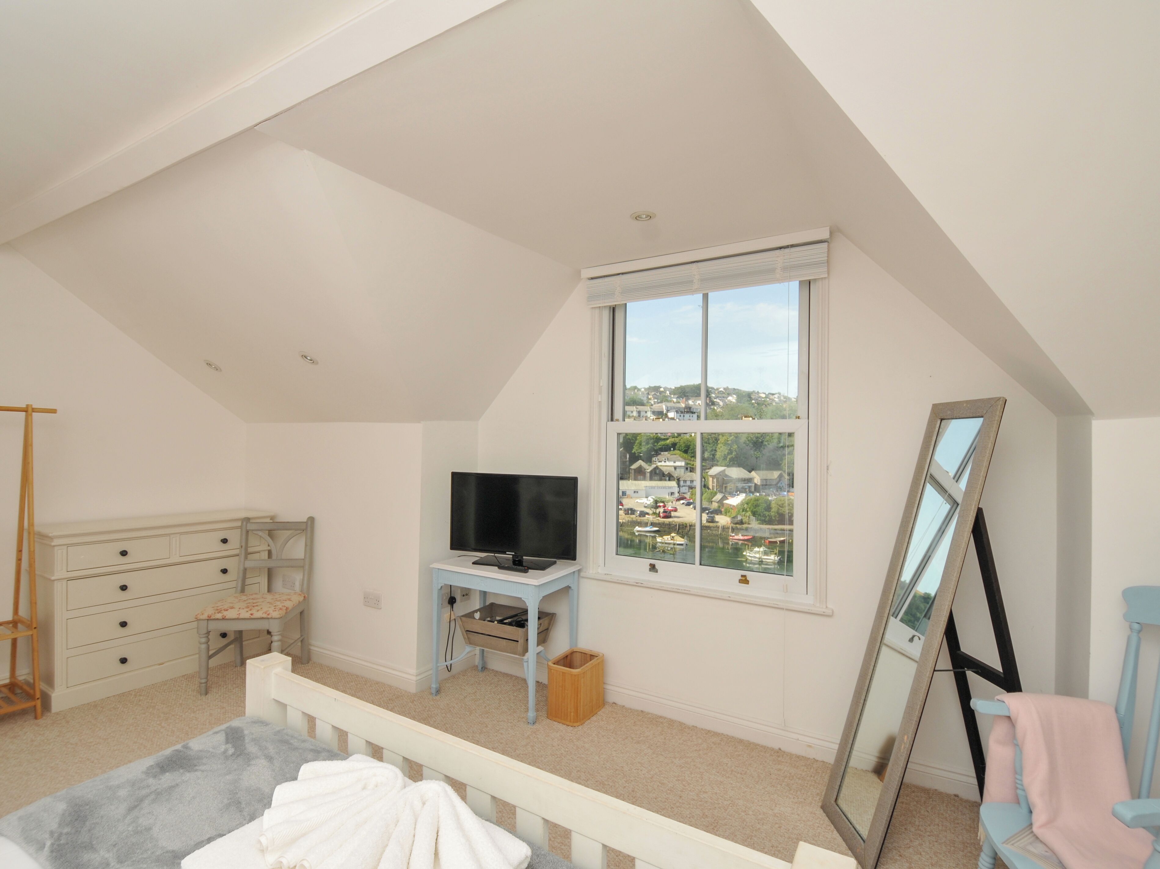 A bedroom with a sloped ceiling, a television on a stand, a dresser, a window with a view of a town, and a mirror leaning against the wall.