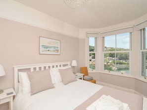 A bedroom with a large bed, a nightstand, a lamp, a chair, and a window with a view of a body of water and buildings. - Overdale (Looe)