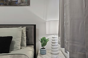 Ein ordentlich eingerichtetes Schlafzimmer mit einem Bett, einem Nachttisch mit Lampe und Pflanze sowie einem Gemälde an der Wand.