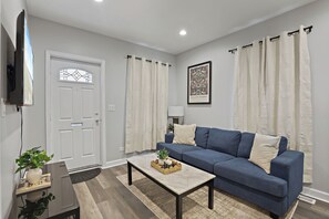Ein Wohnzimmer mit einem blauen Sofa, einem Couchtisch mit dekorativem Korb, einer weißen Tür mit Glasfenster und einem gerahmten Bild an der Wand.