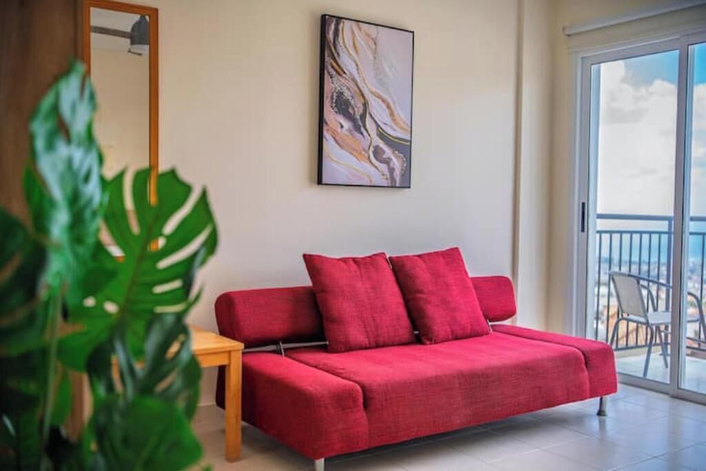 Un divano rosso con cuscini, un tavolino in legno, un grande quadro astratto e un balcone con vista.