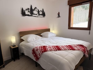 A neatly made bed with a white comforter and a red patterned throw blanket, a nightstand with a lamp, a wall-mounted decoration of a mountain scene, and a window with a view of trees. - Balcony, tv, 30m², Pralognan-la-Vanoise (Pralognan-la-Vanoise)