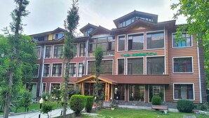 Um hotel com vários andares, entrada central, gramado bem cuidado e área externa com mesas e cadeiras.