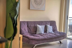 A purple sofa with cushions, a wooden chair, a large green plant, and a wall-mounted circular artwork. - Peyia Pearl 31 – Elevated Sea View Studio (Pegeia)