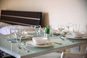 Une table à manger élégamment dressée, avec de la vaisselle en verre transparent, des assiettes et des couverts, une petite plante en pot, et un lit avec une tête de lit sombre en arrière-plan.