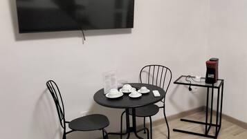 Una stanza con una TV a schermo piatto montata a parete, un tavolo rotondo con sedie, una macchina per il caffè e un tavolino con tazze e piattini.