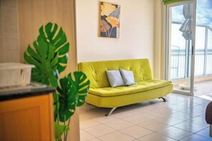A bright yellow sofa with two pillows, a large green plant, and a painting on the wall. - Peyia Pearl 27 – Sea View Studio w/ Large Veranda (Pegeia)