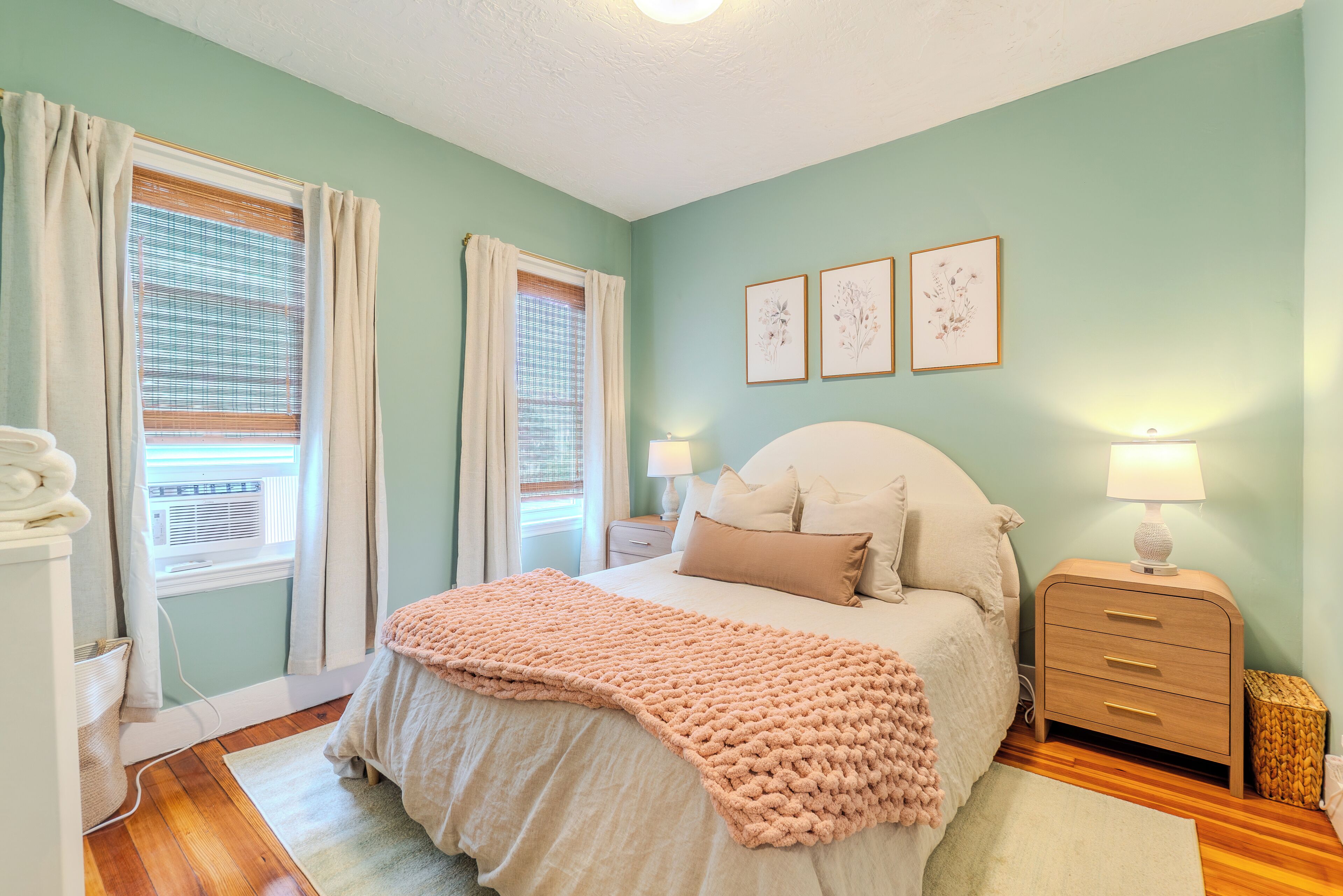 A bedroom with a bed, bedside tables, a nightstand, a window with curtains, and wall art.