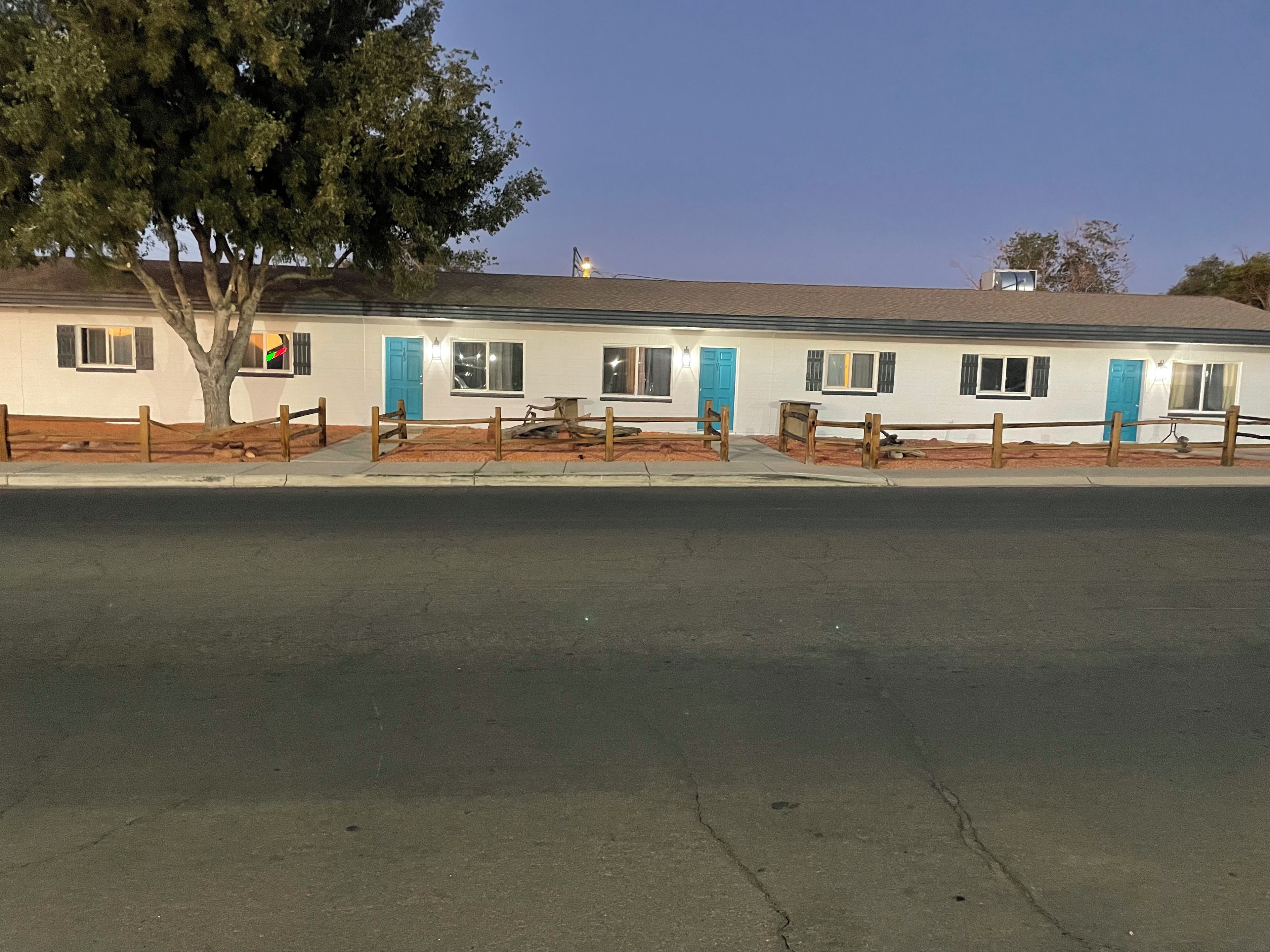 A row of single-story motel buildings with individual doors and windows, a tree in front, and a paved road in the foreground.