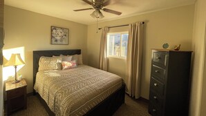 A bedroom with a bed, a ceiling fan, a nightstand with a lamp, a dresser, and a window with curtains.