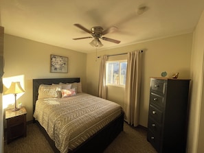 A bedroom with a bed, a ceiling fan, a nightstand with a lamp, a dresser, and a window with curtains.