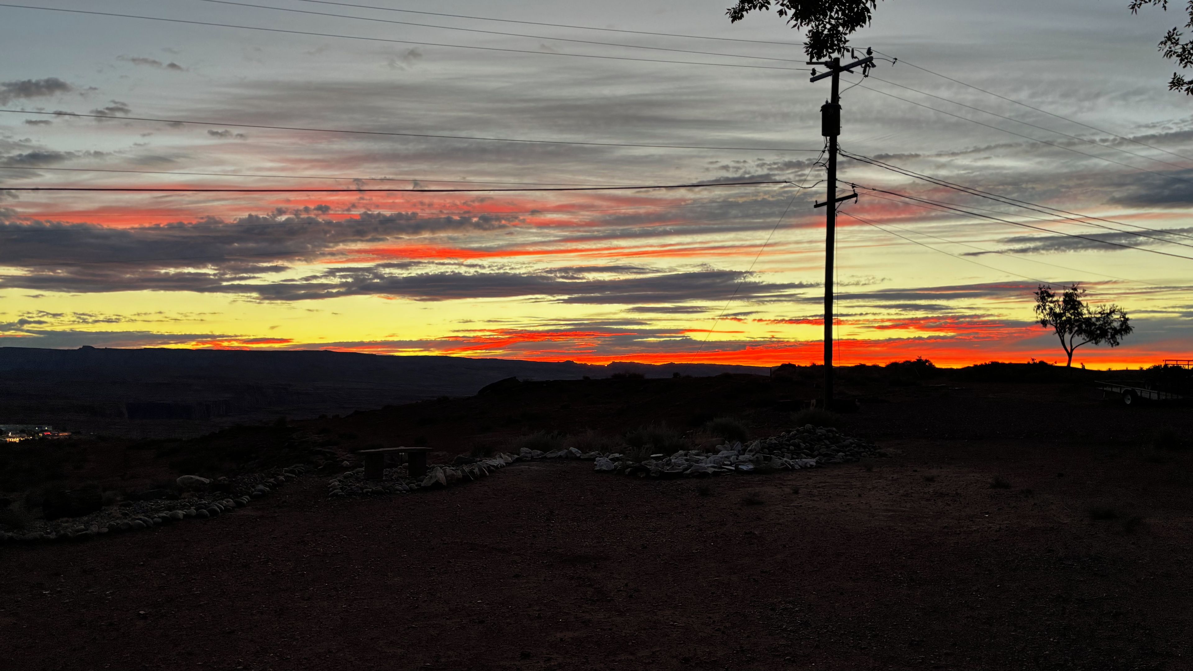 A sunset over a hilly landscape with a dirt road, a utility pole, and sparse vegetation.