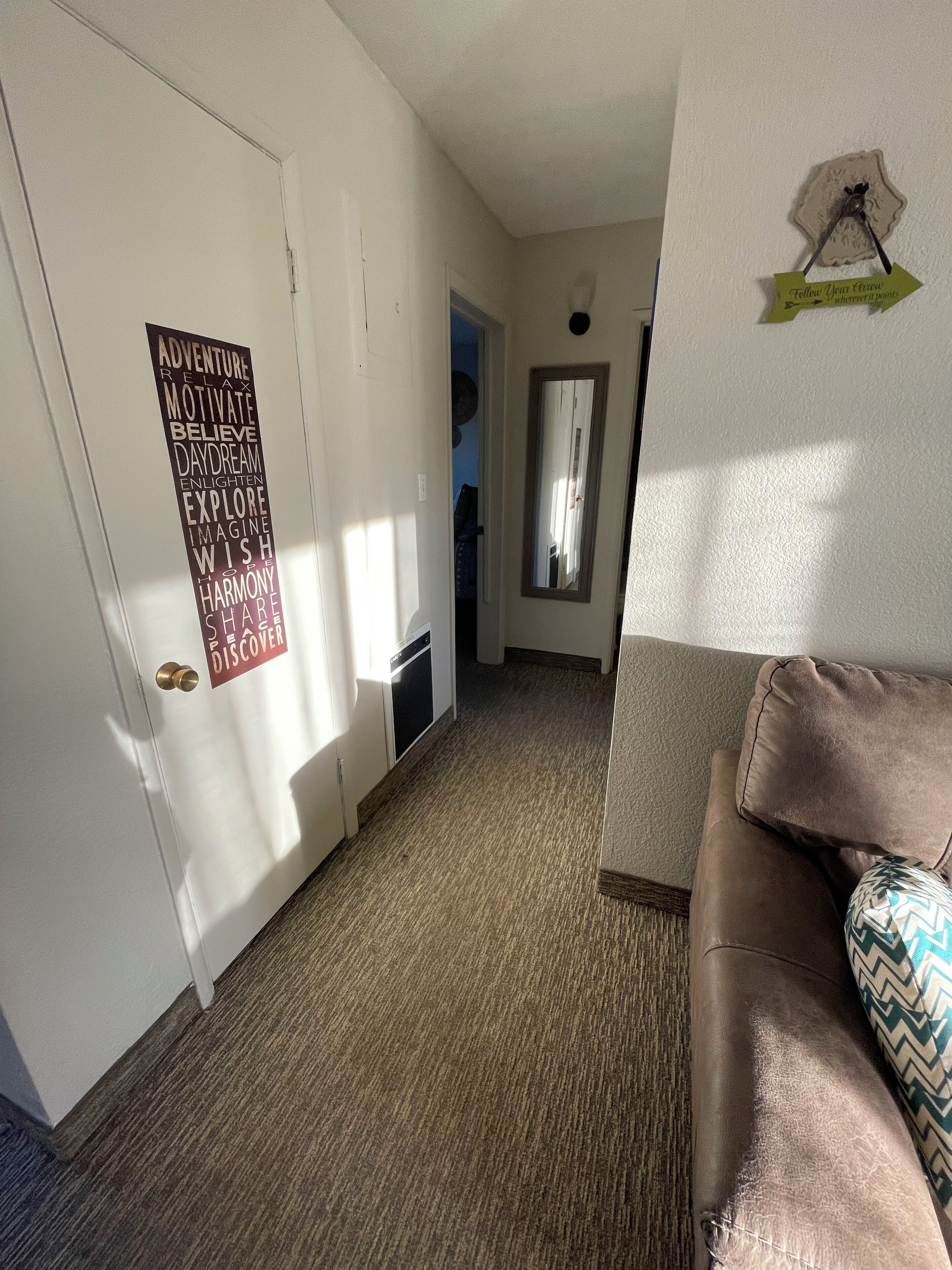 A hallway with a carpeted floor, a door with a motivational poster, a mirror, and a couch with patterned cushions.