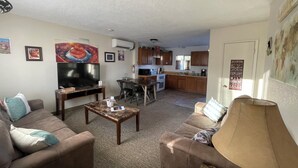 A living room with a TV, sofa, coffee table, and a kitchen in the background.
