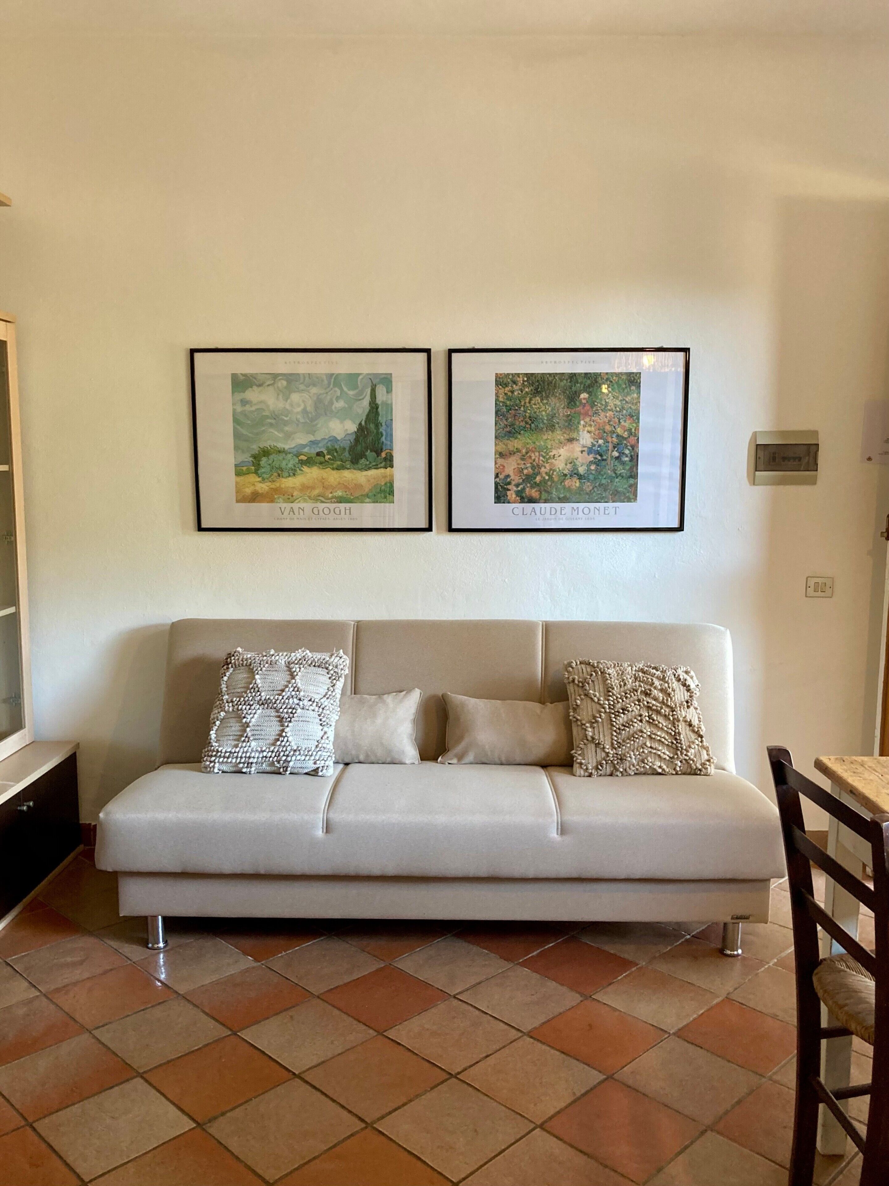 Un salon avec un canapé beige, deux œuvres d’art encadrées et un sol carrelé.