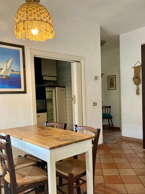 Un coin repas avec une table et des chaises en bois, un luminaire suspendu, un tableau encadré et une cuisine visible à travers une porte ouverte.