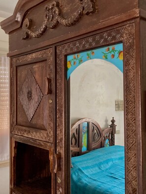 Une armoire en bois ornée de sculptures complexes, avec un miroir qui reflète un lit recouvert d’une couverture bleue.