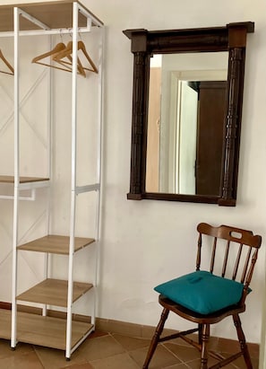 A room with a white metal shelving unit, a wooden chair with a teal cushion, and a dark wooden mirror on the wall.