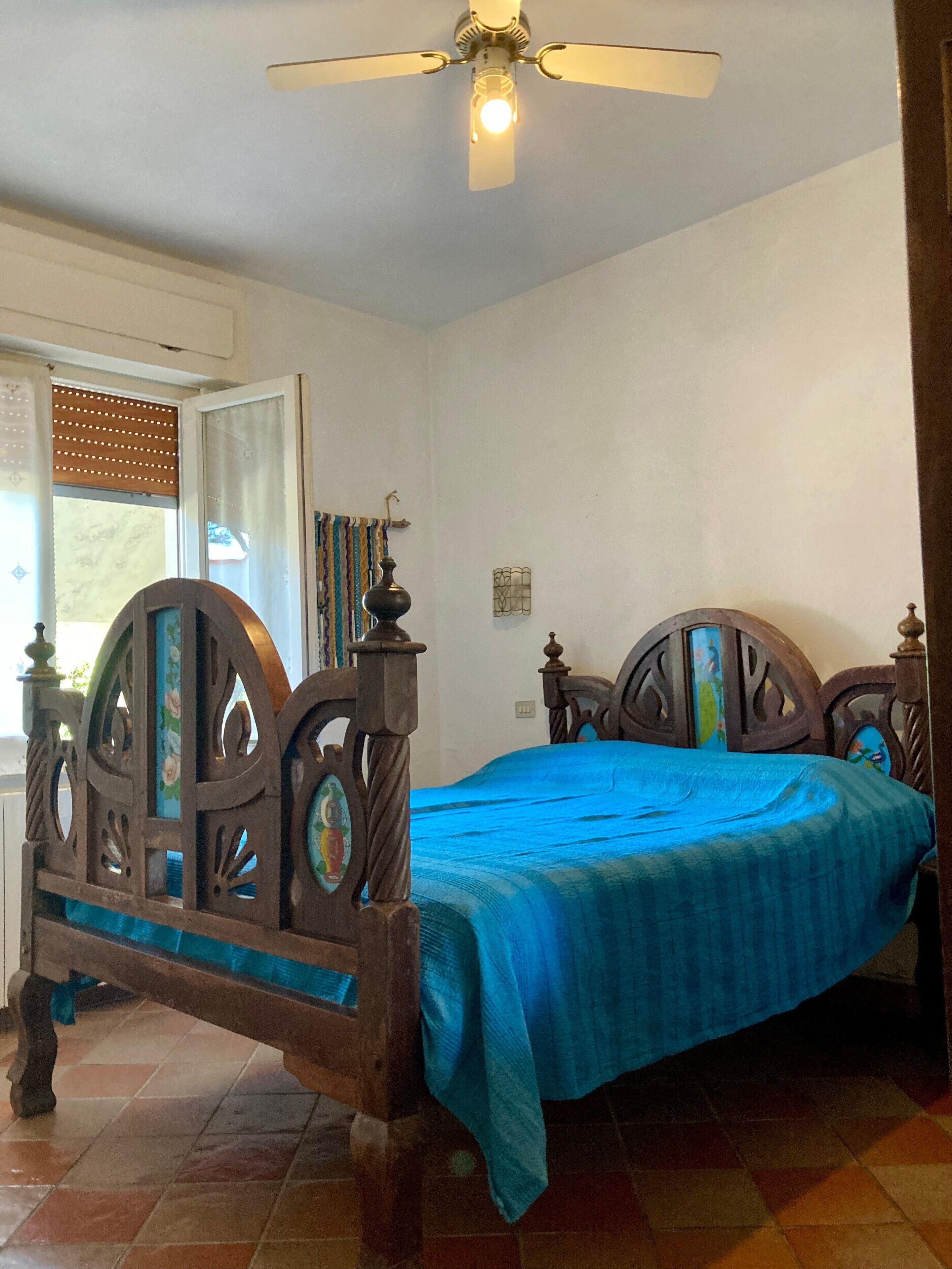 Une chambre avec un lit en bois, un ventilateur de plafond et une fenêtre avec des stores.