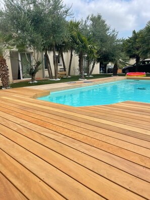 A wooden deck surrounding a swimming pool with trees and a building in the background. - Vrbo Property (Lagord)