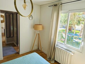Una habitación con cama, una lámpara de pie, una ventana con cortinas, un radiador y vistas a un jardín exterior.
