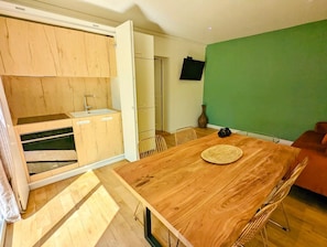 A compact kitchen with wooden cabinets, a built-in oven, and a dining area with a wooden table and chairs. - Vrbo Property (Lagord)