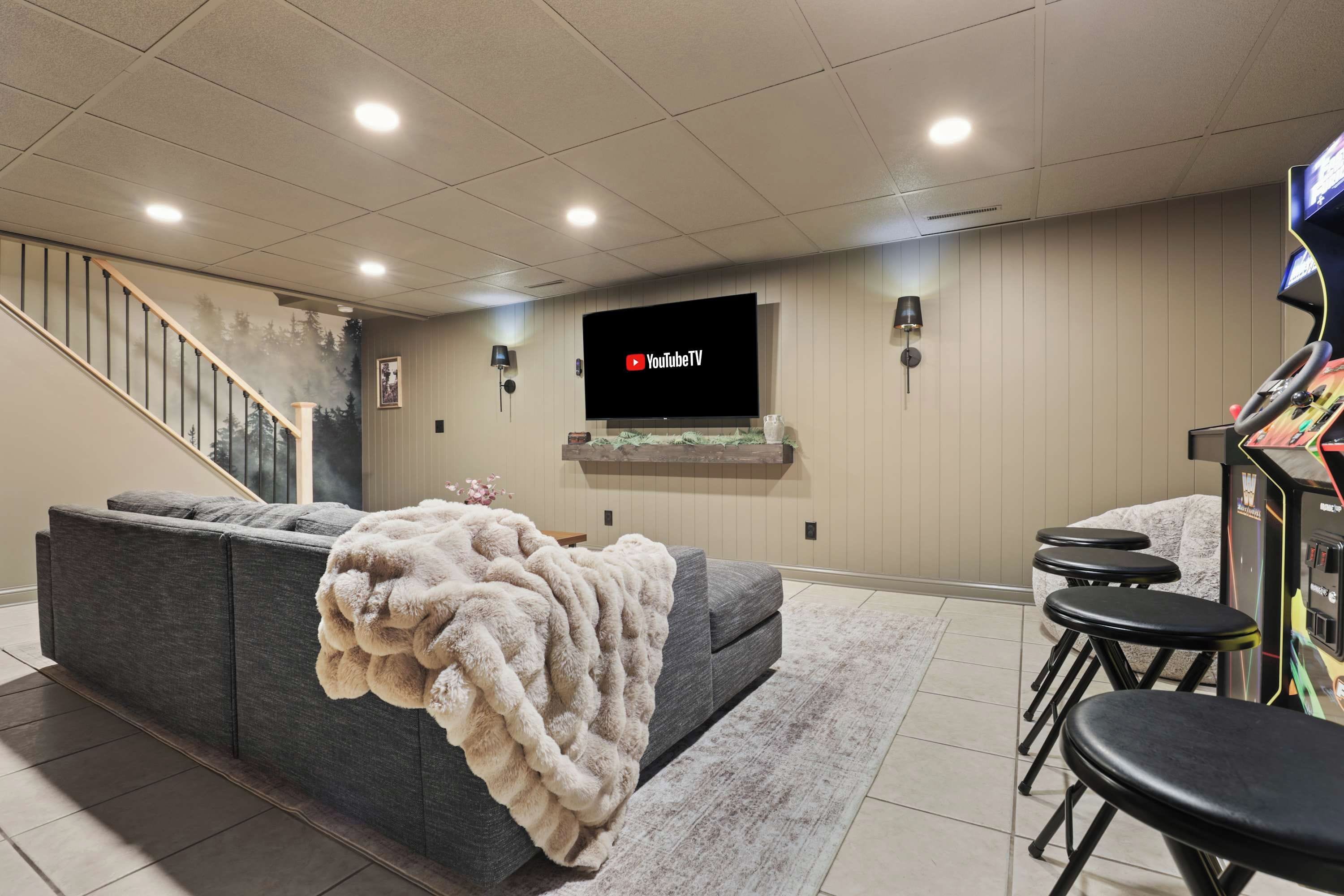 Een moderne woonkamer met een grijze hoekbank, een flatscreen televisie aan de muur, een trap en een speelgedeelte met krukken en een race stuurwiel.