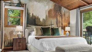 A bedroom with a large bed, bedside lamps, a nightstand, and a view of trees through the window.