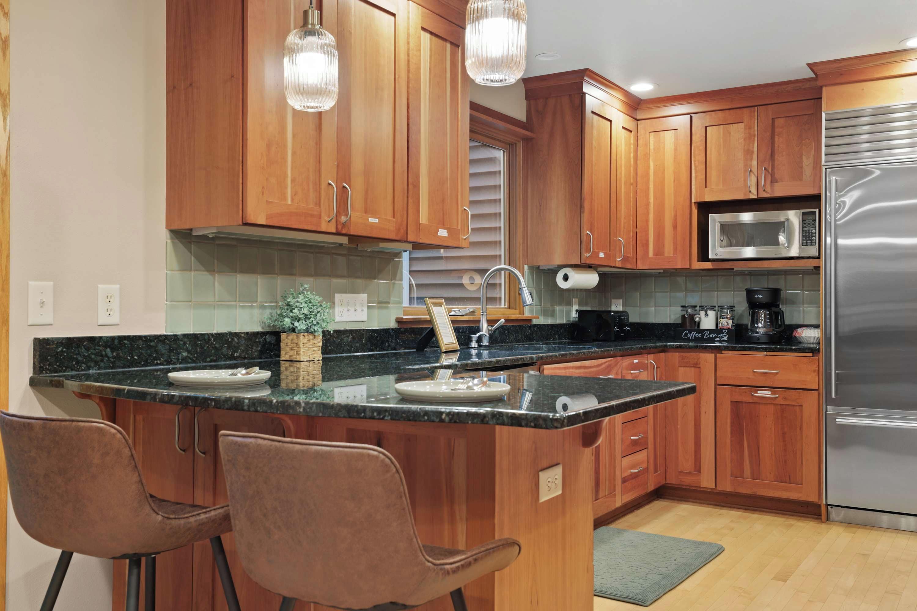 A kitchen with wooden cabinets, a stainless steel refrigerator, a microwave, and a coffee maker. There are two bar stools and a small potted plant on the counter.