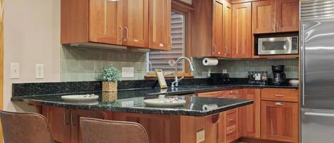 A kitchen with wooden cabinets, a stainless steel refrigerator, a microwave, and a coffee maker. There are two bar stools and a small potted plant on the counter.