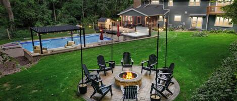A backyard with a circular fire pit surrounded by chairs, a swimming pool, and a covered patio area.