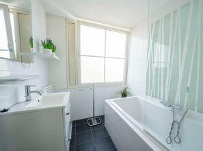 Une salle de bain avec un lavabo blanc, un miroir et une baignoire. Il y a une fenêtre donnant sur l’extérieur.