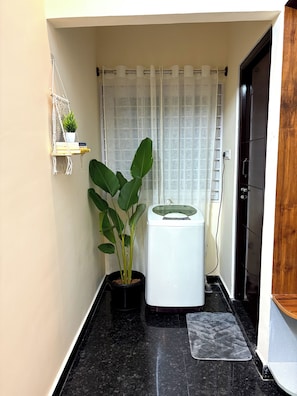 A compact bathroom with a potted plant, a white washing machine, and a shower curtain. - Luxury Stay 20 mins away from Airport! (Bengaluru)