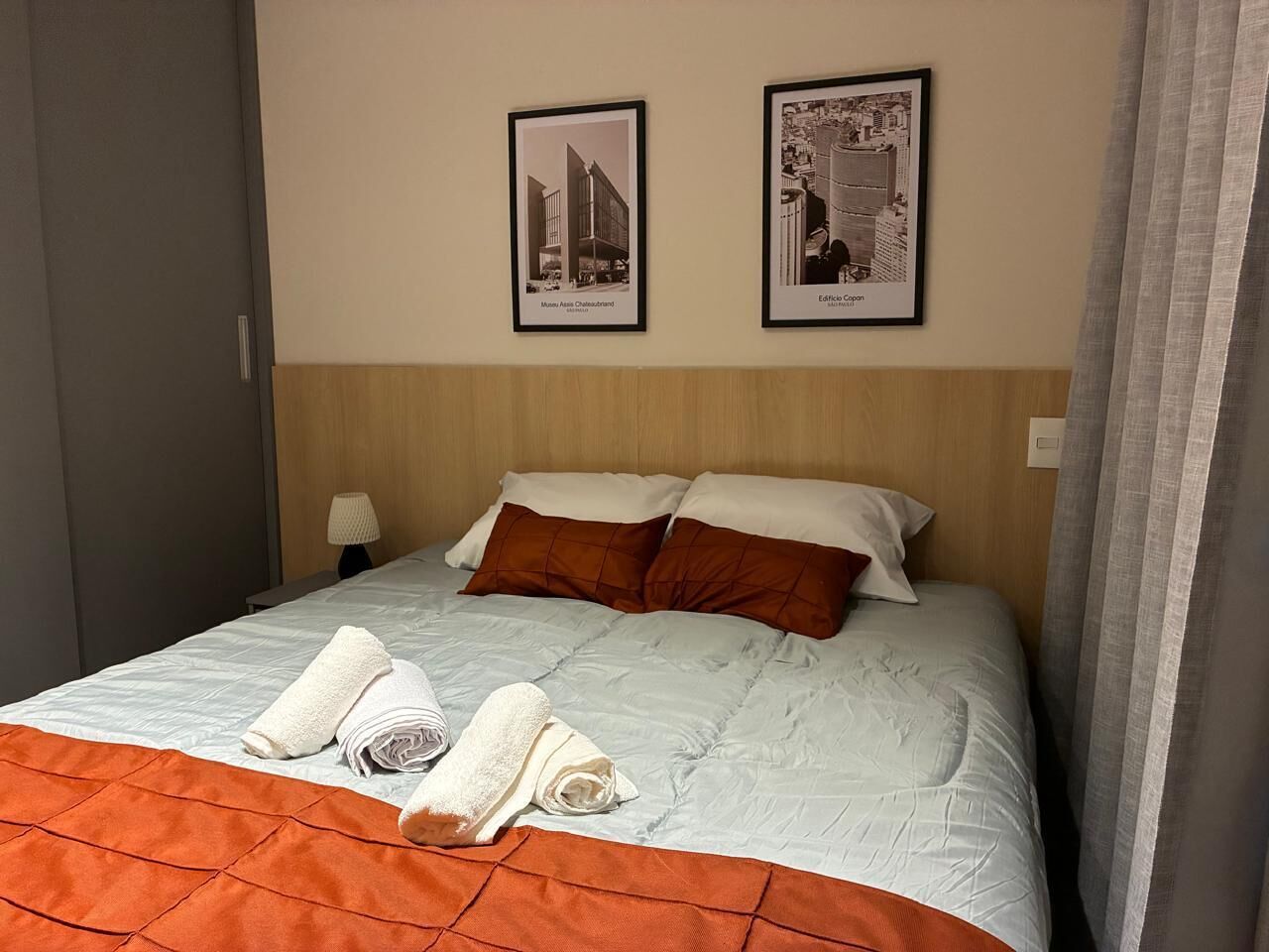 A hotel room with a bed, pillows, a folded towel, and two framed pictures on the wall.