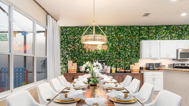A dining area with a long wooden table set for a meal, surrounded by white chairs, with a neon sign and a green leafy wall in the background. - Disney Themed Resort Home With Lake View (Kissimmee)
