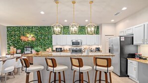 A modern kitchen with white cabinets, a central island with bar stools, a neon sign, and a green leafy backsplash. - Disney Themed Resort Home With Lake View (Kissimmee)