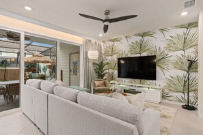 Un salon moderne avec un canapé, un téléviseur, un ventilateur de plafond et un papier peint aux motifs tropicaux.