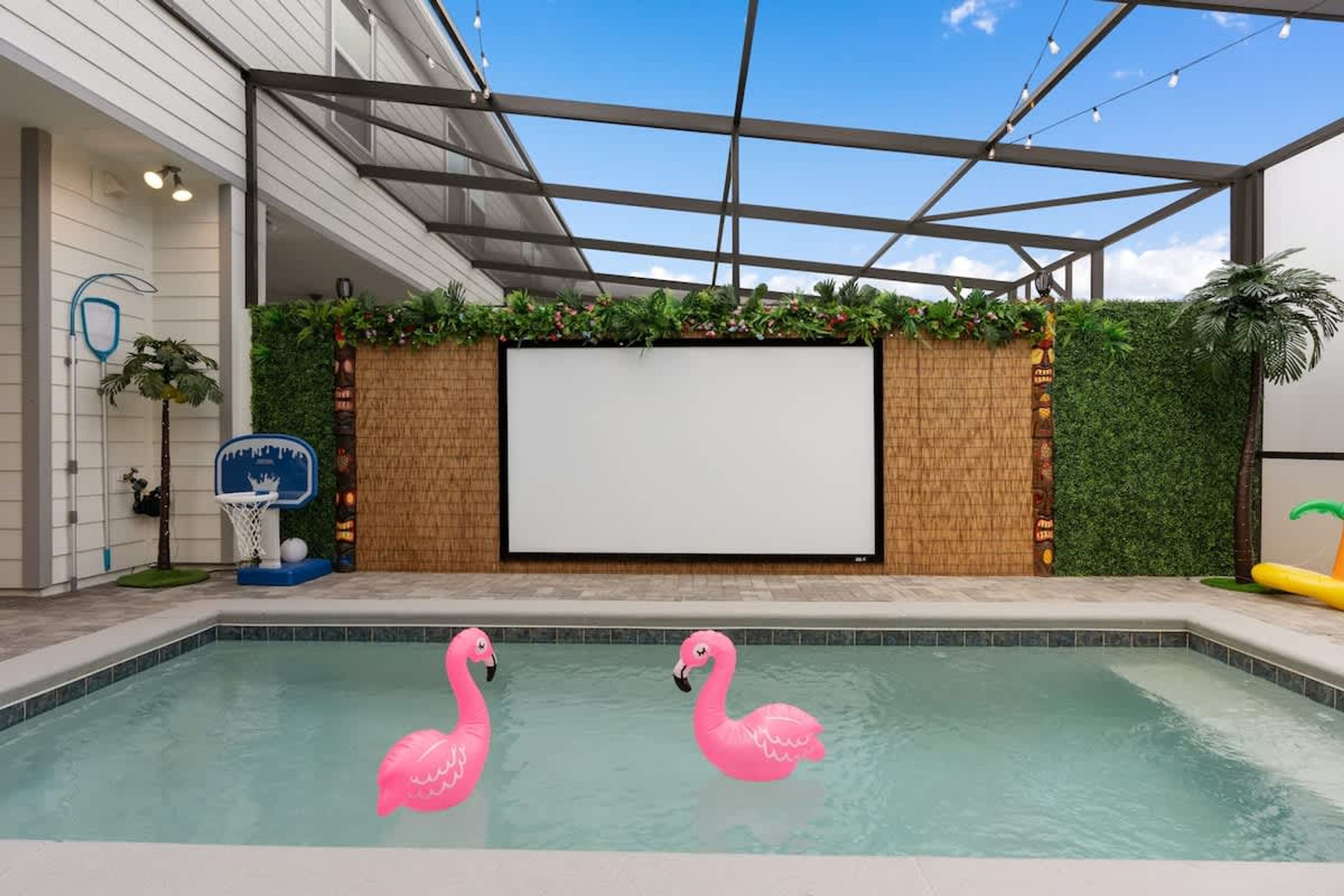 A swimming pool with pink flamingo floats, a basketball hoop, and a large blank screen on a wall.