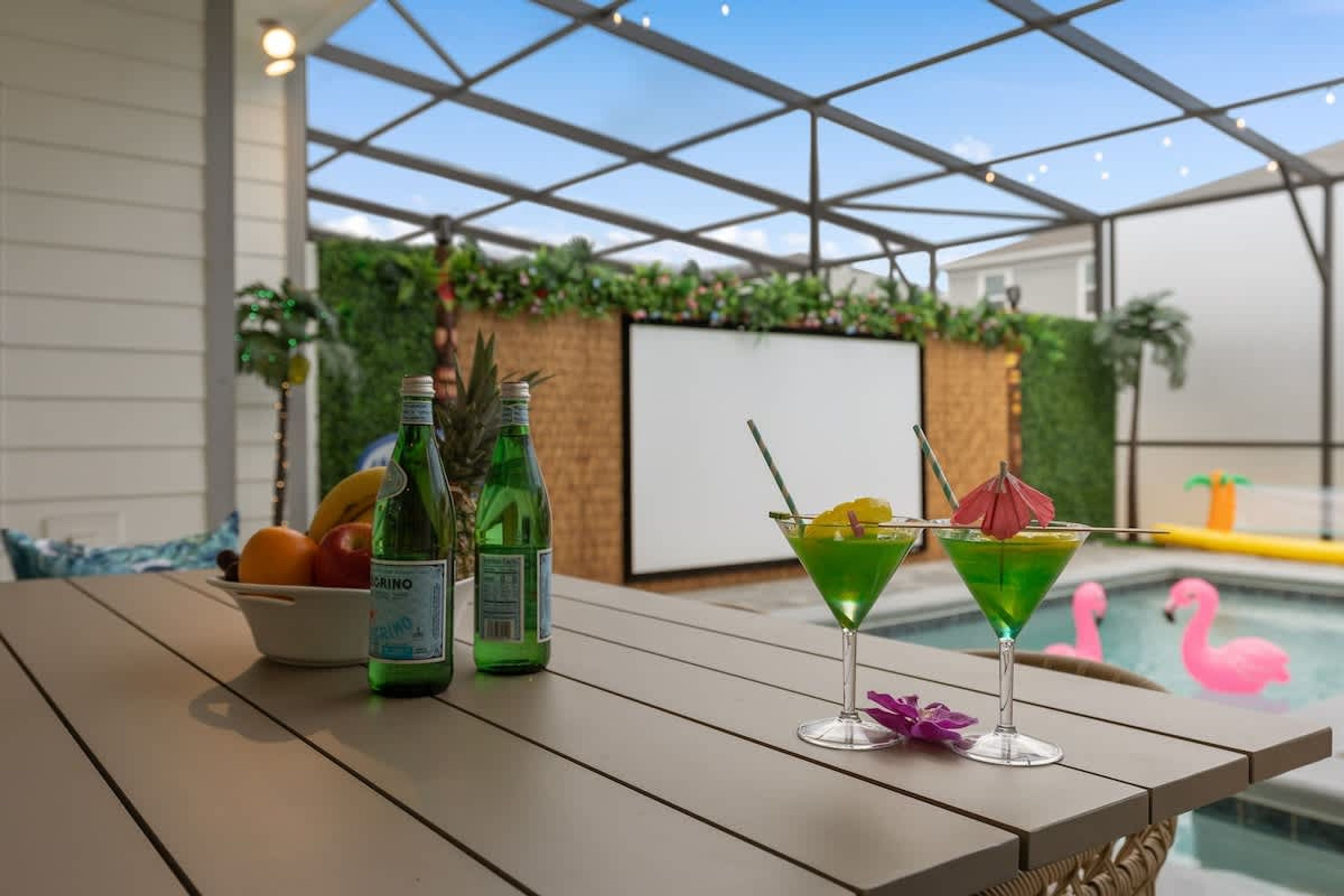 A poolside table with two bottles and two martini glasses, a bowl of fruit, and a pool with pink flamingos.