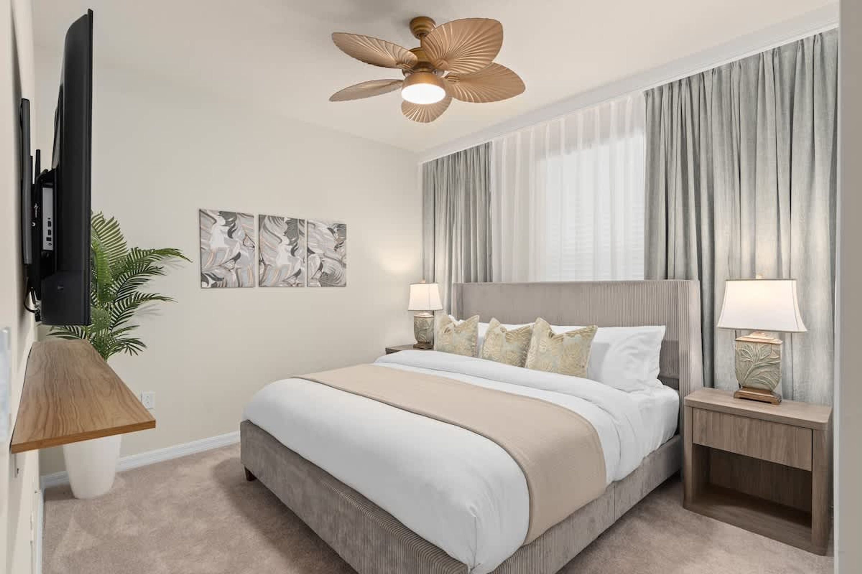 Habitación de hotel con cama, ventilador de techo, televisión, obras de arte en la pared y cortinas.