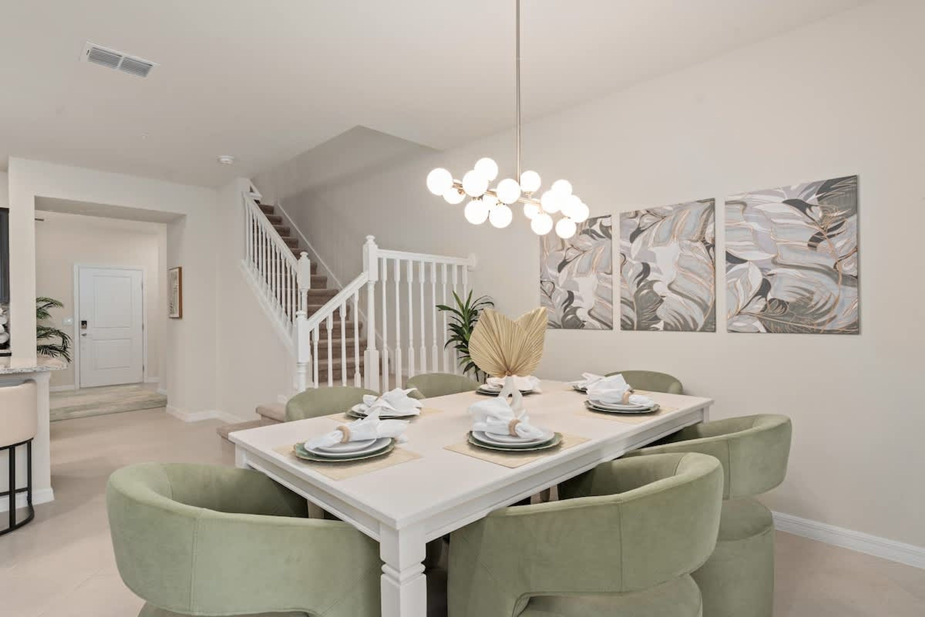 Un coin repas avec une table blanche dressée pour quatre personnes, des chaises à revêtement vert, un escalier blanc et des œuvres d’art abstraites accrochées au mur.